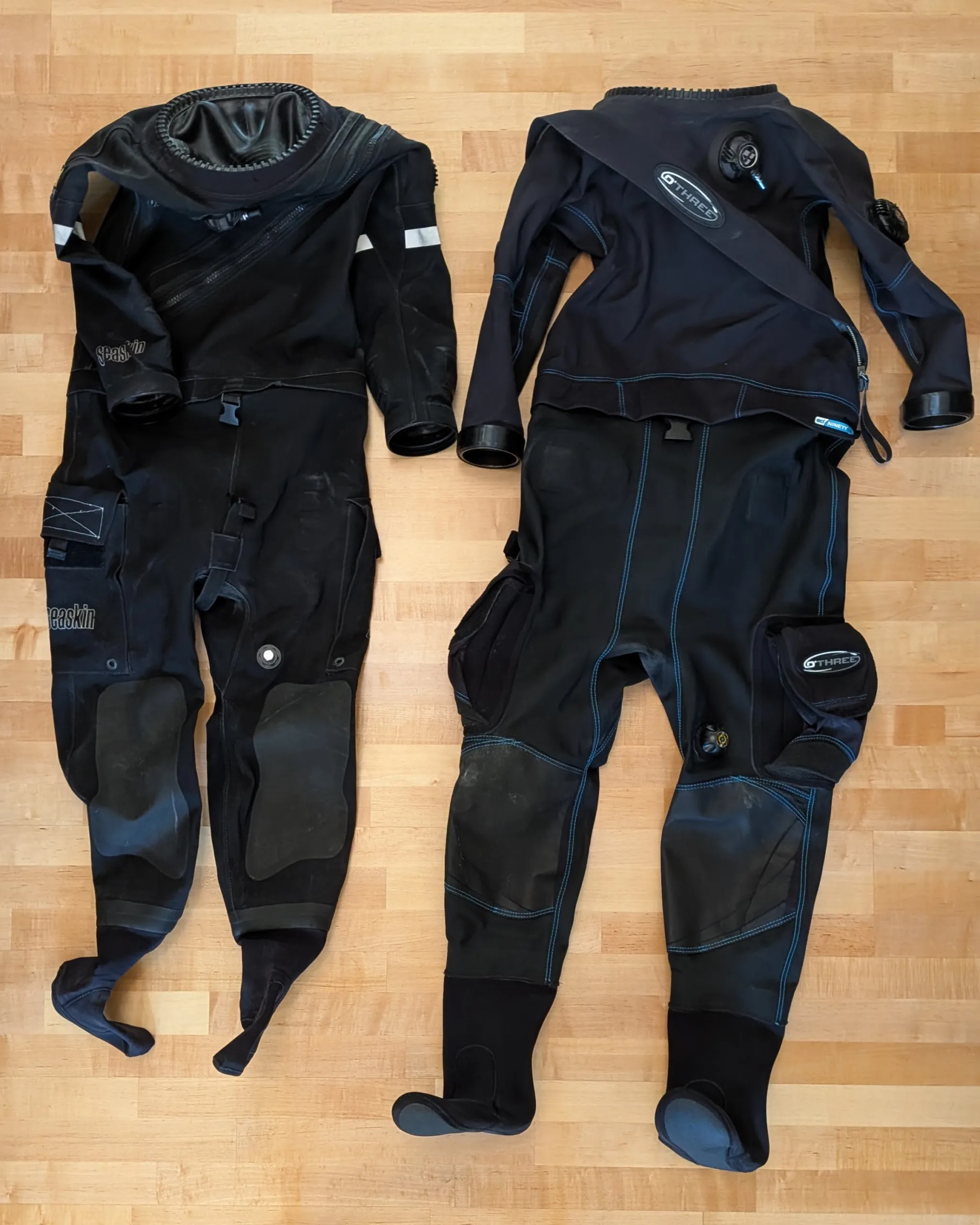 Seaskin Nova and O'Three 90Ninety Hybrid drysuits laid flat side by side on wood floor